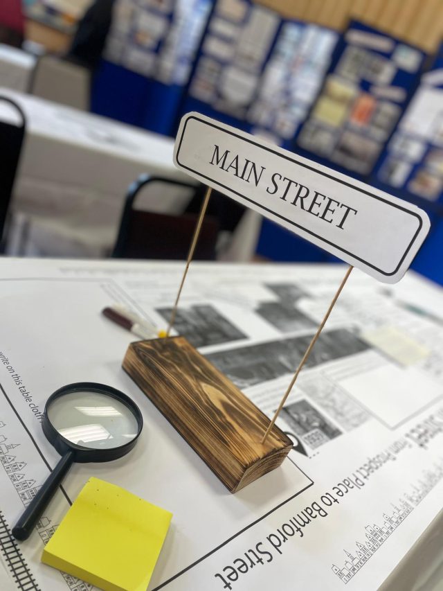 a street sign for Main street sits upon a table with photos and maps. In the background is a visual display of photos and information